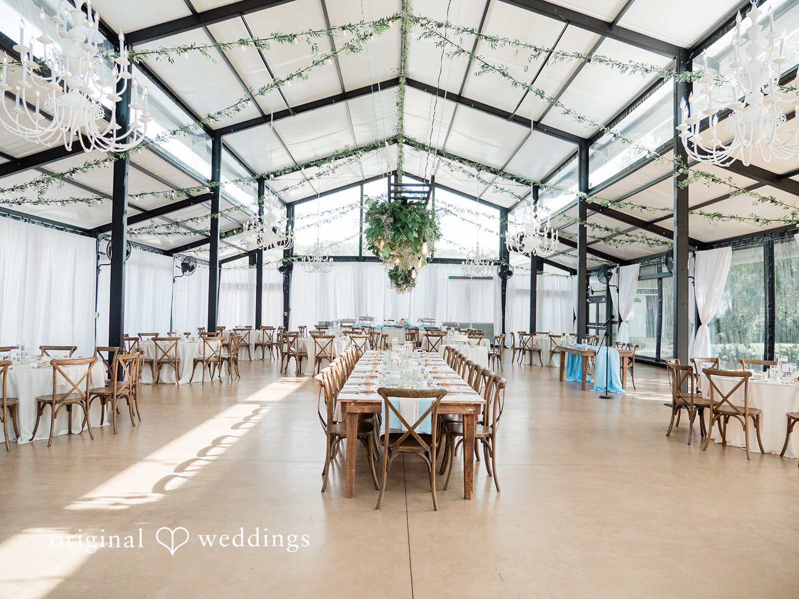 Paige + Stephen An empty wedding reception hall with long wooden tables and a high, modern glass-paneled ceiling structure.