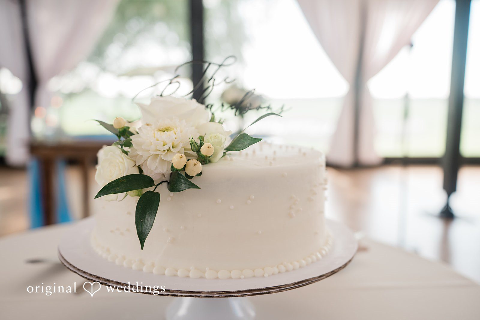Paige + Stephen A minimalist two-tiered white wedding cake decorated with elegant white roses and green foliage.
