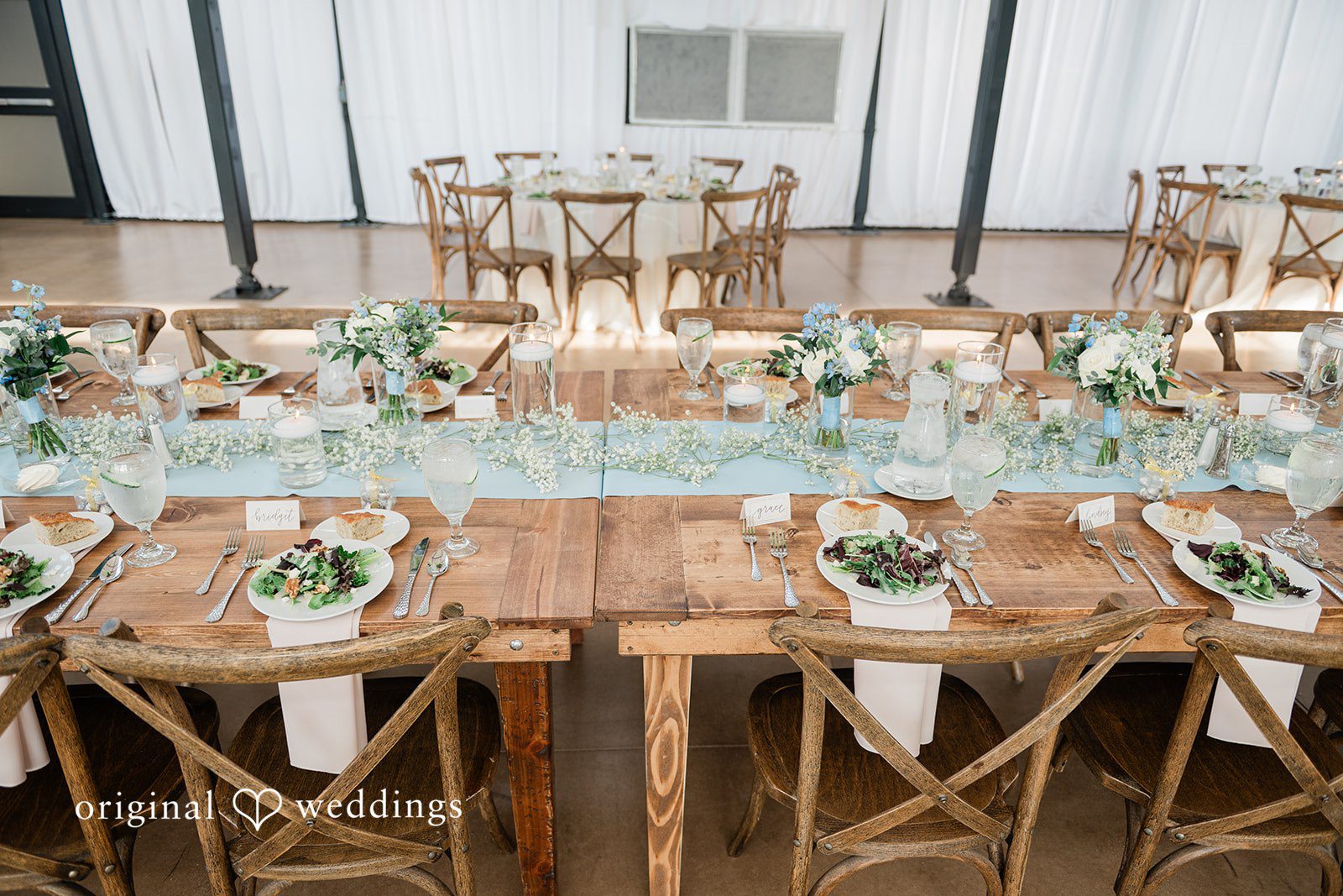 Paige + Stephen A wooden banquet table set with white plates, blue napkins, glass goblets, and eucalyptus greenery.