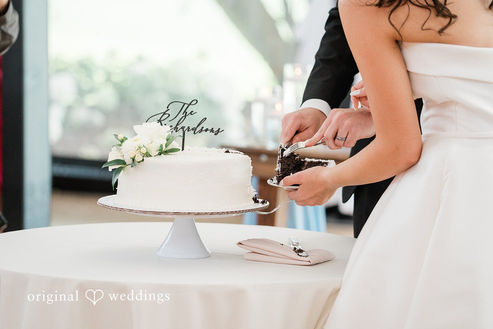 Paige + Stephen A close-up view of a bride cutting a tiered white wedding cake at the reception table.