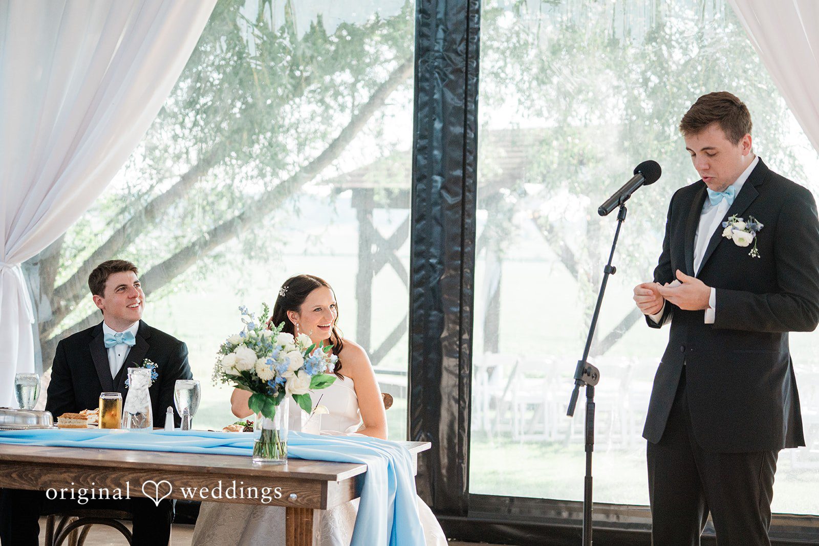 Paige + Stephen A man in a black suit and blue bowtie speaks into a microphone while a bride and another man look on and smile from a nearby table.
