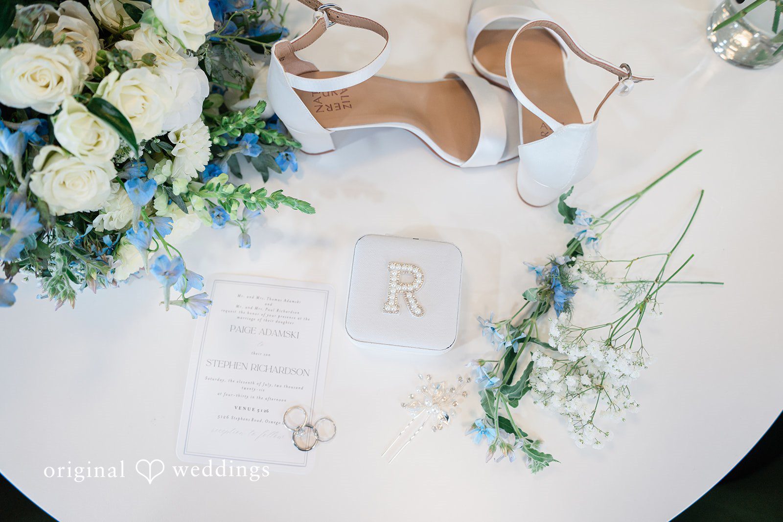 Paige + Stephen A pair of white high-heeled bridal shoes surrounded by white flowers and delicate green stems.