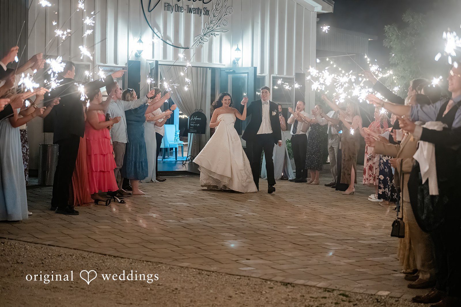 Paige + Stephen A bride and groom walking together at night surrounded by guests holding glowing sparklers.