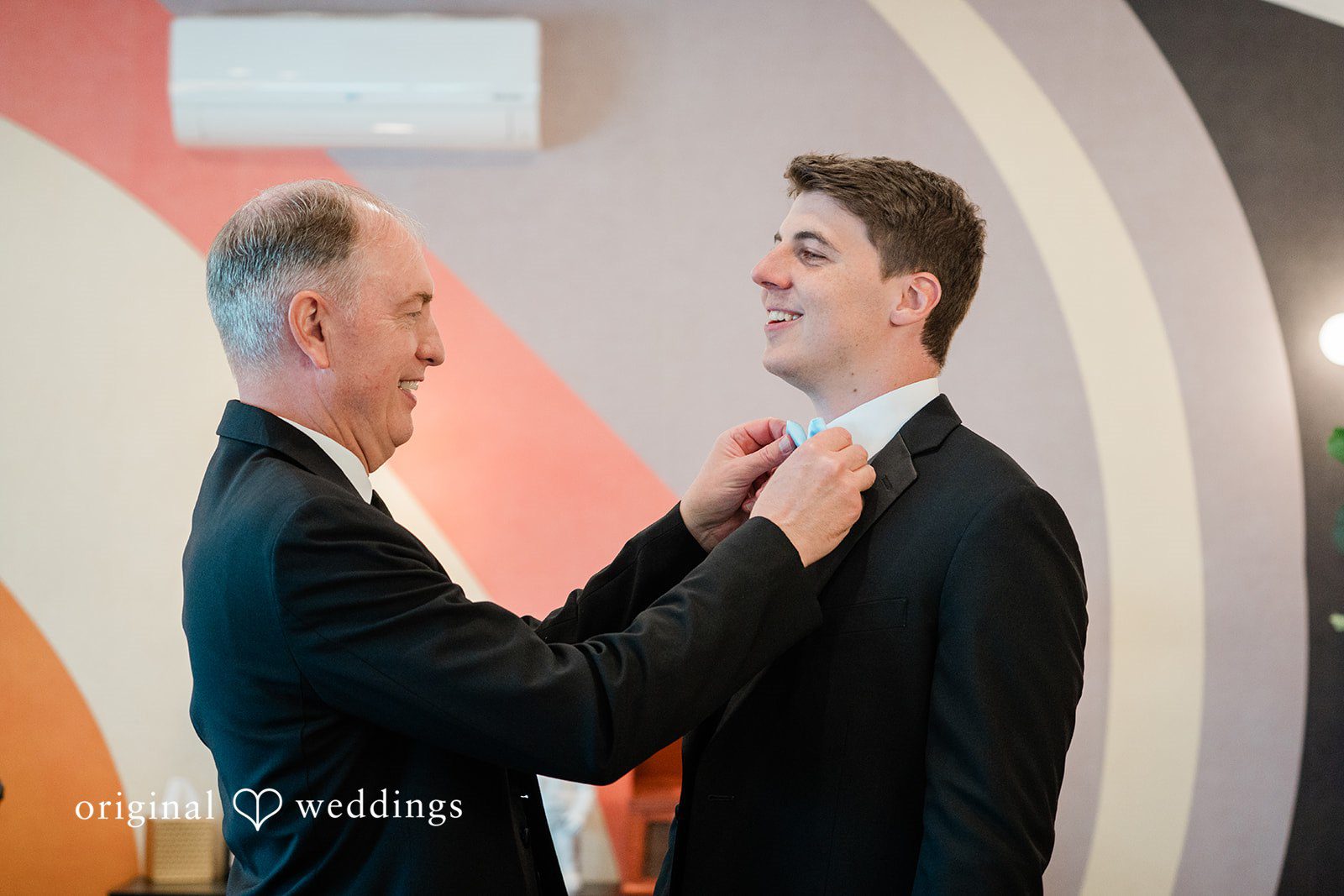 Paige + Stephen A groom in a black suit adjusting a man's tie as they look at each other and smile.