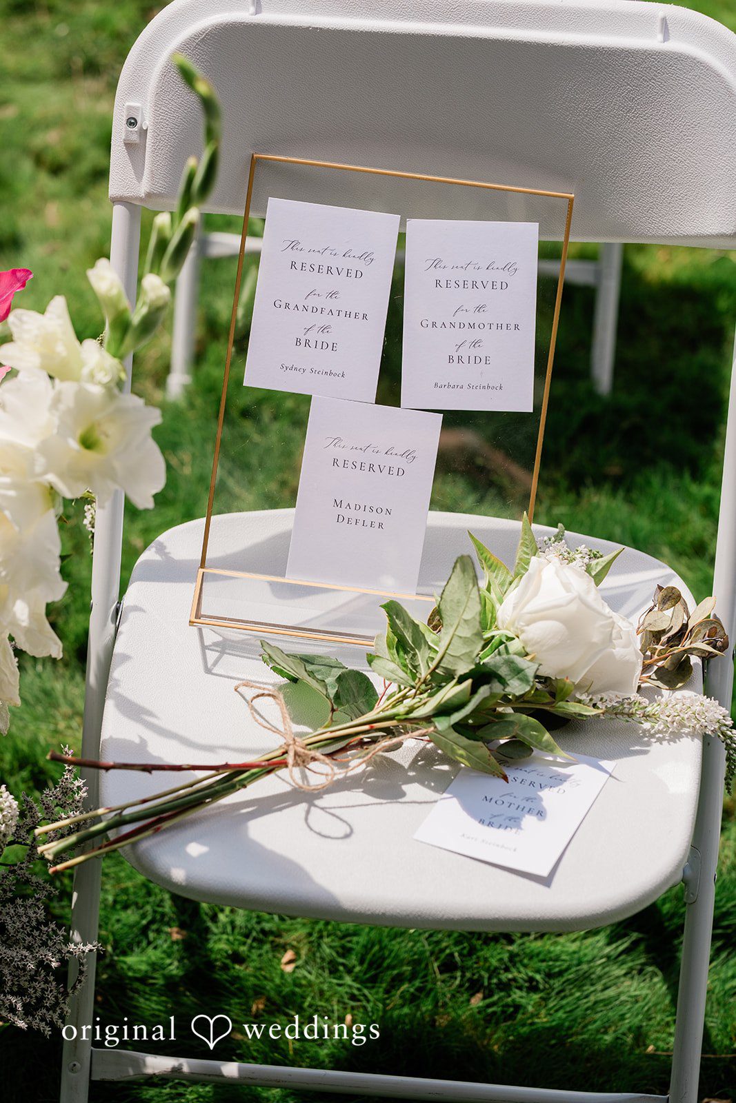 A white reception sign or seating chart displayed on an easel surrounded by greenery.
