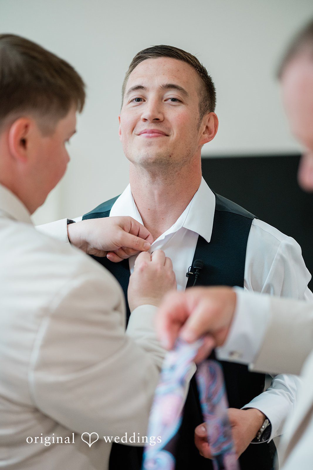 A groom smiling while getting his tie adjusted by someone before the wedding.