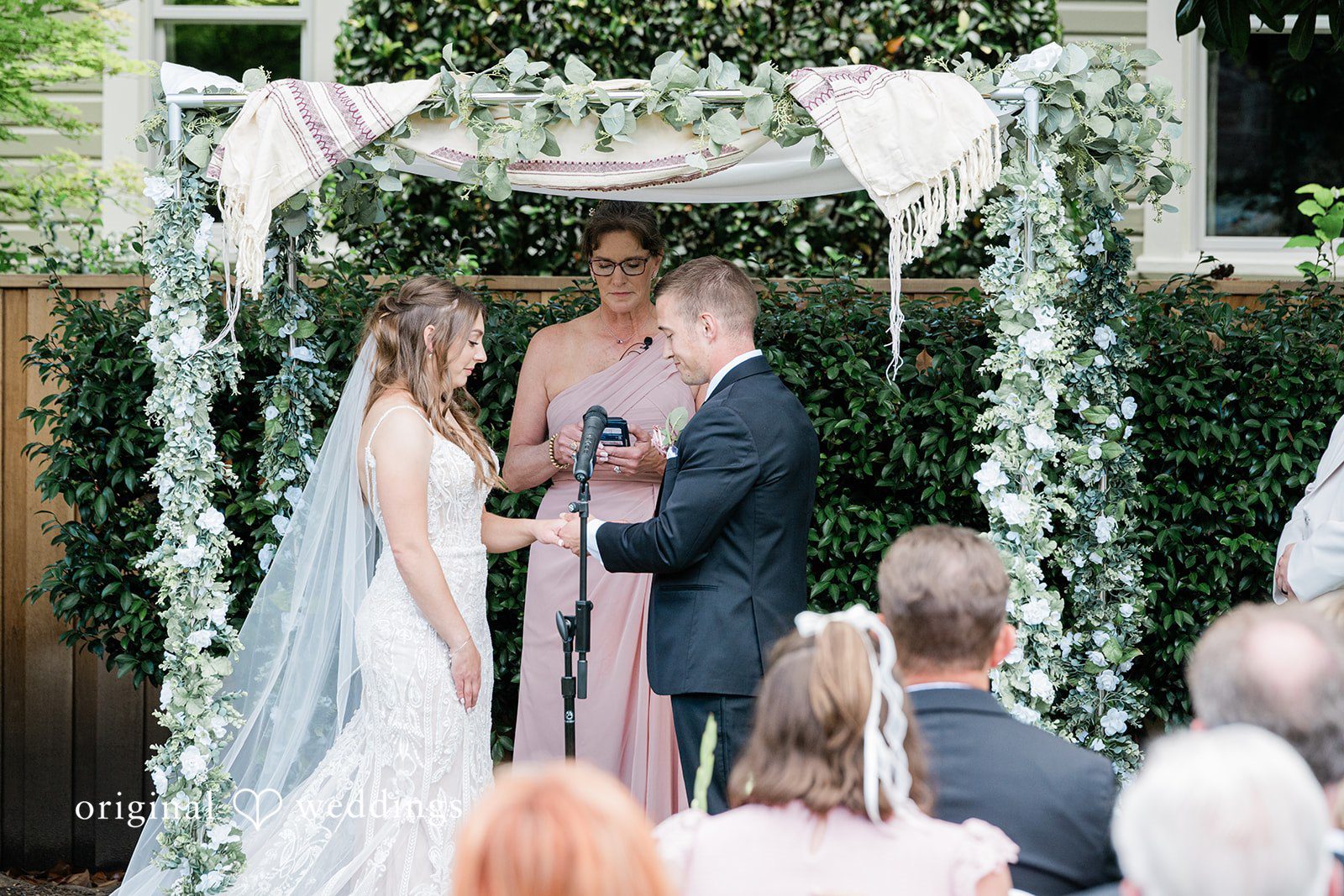 A bride and groom standing under an arch decorated with white fabrics and greenery.