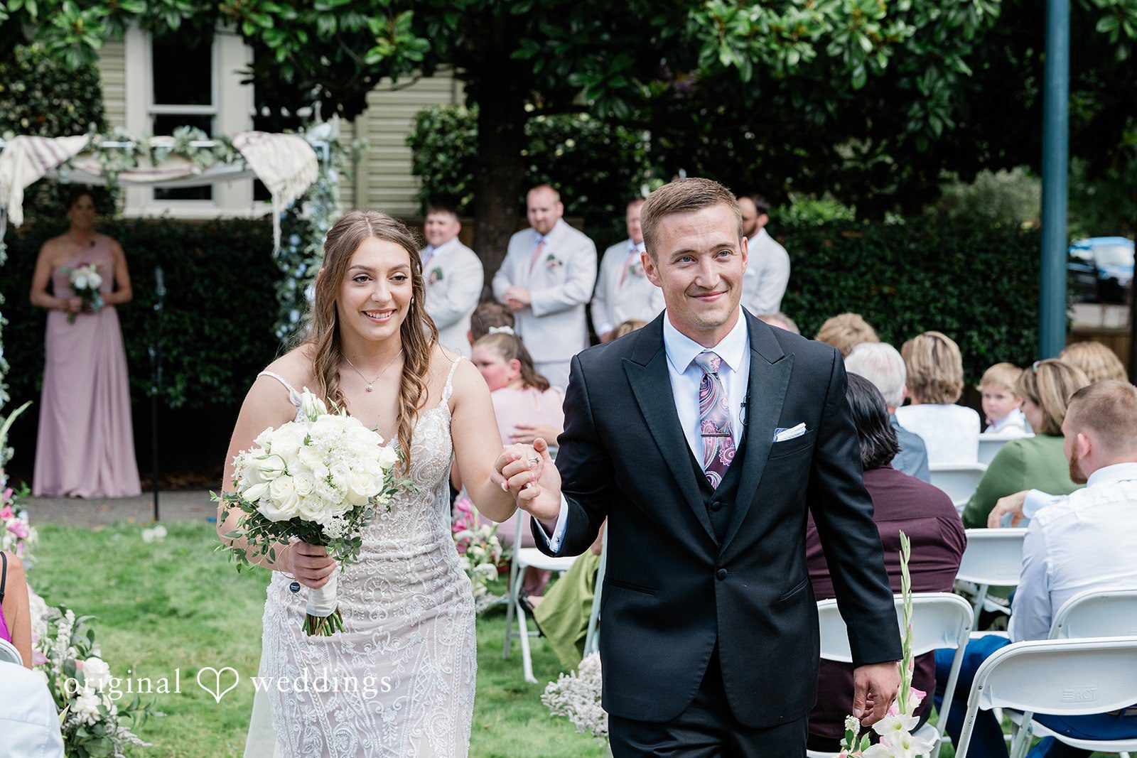 A bride and groom walking up a brick path together while guests smile in the background.