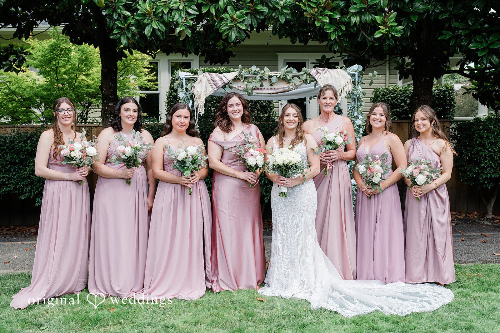 A bridal party posing together on a grassy lawn with bridesmaids dressed in long pink gowns.