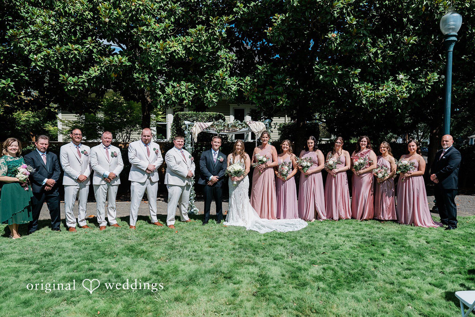 A wedding party arranged in a long line for a group photo in front of dense greenery.