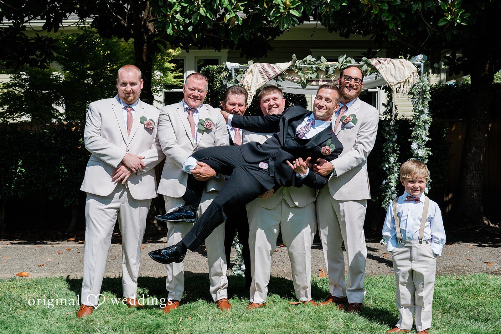 A group of groomsmen playfully lifting the groom up in the air on a pathway.