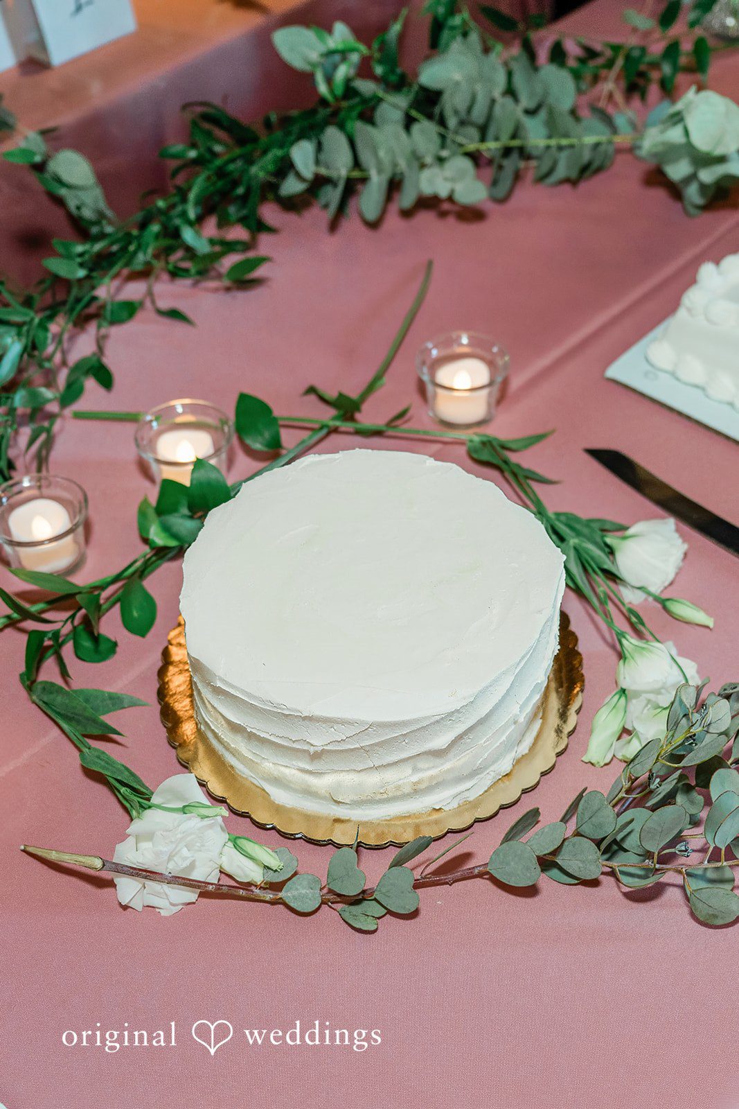 A smooth, single-tier white wedding cake sitting on a mauve-colored table fabric.