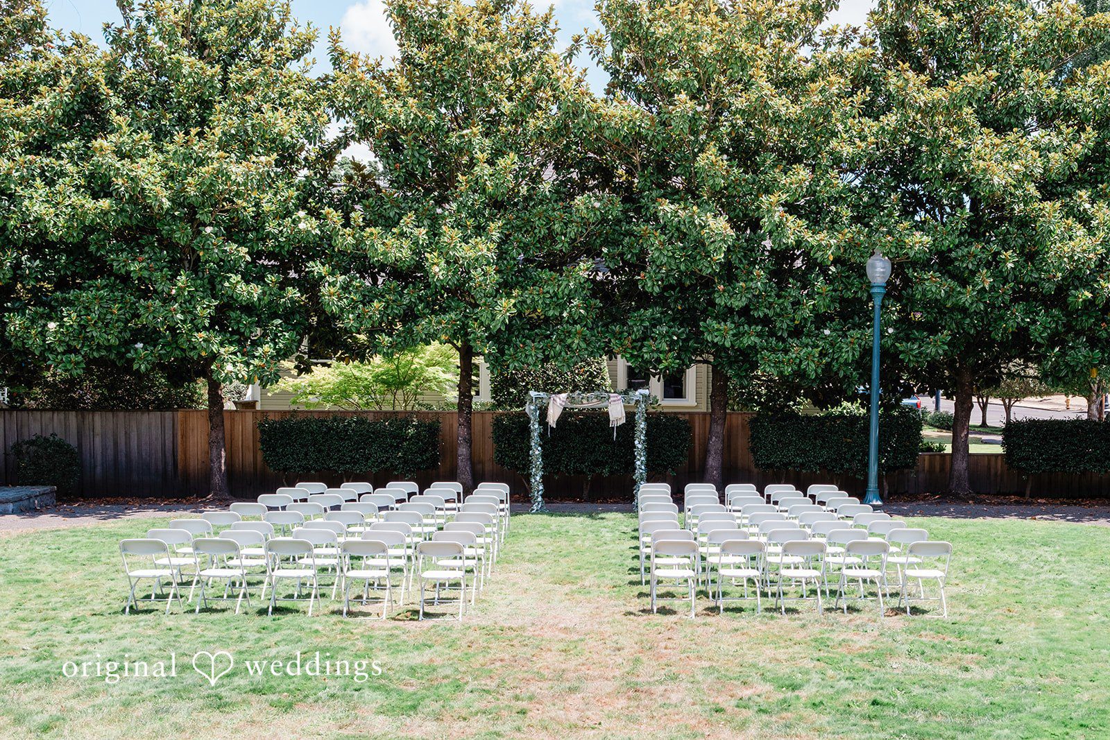 An rows of empty white chairs arranged for an open-air ceremony under tall green trees.