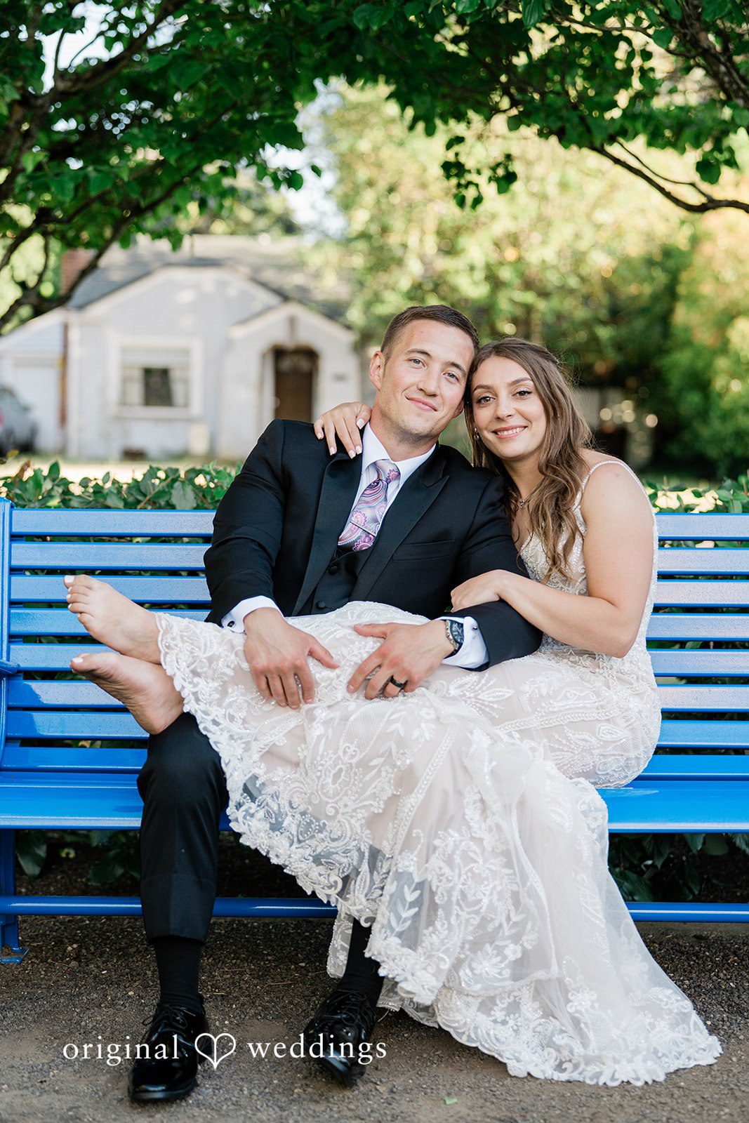 A bride and groom sitting together and smiling on a blue park bench.