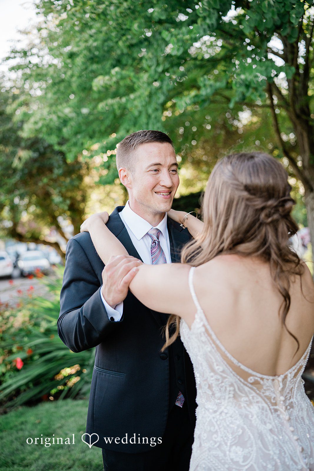 A groom smiling while embracing his bride from behind on a pathway.