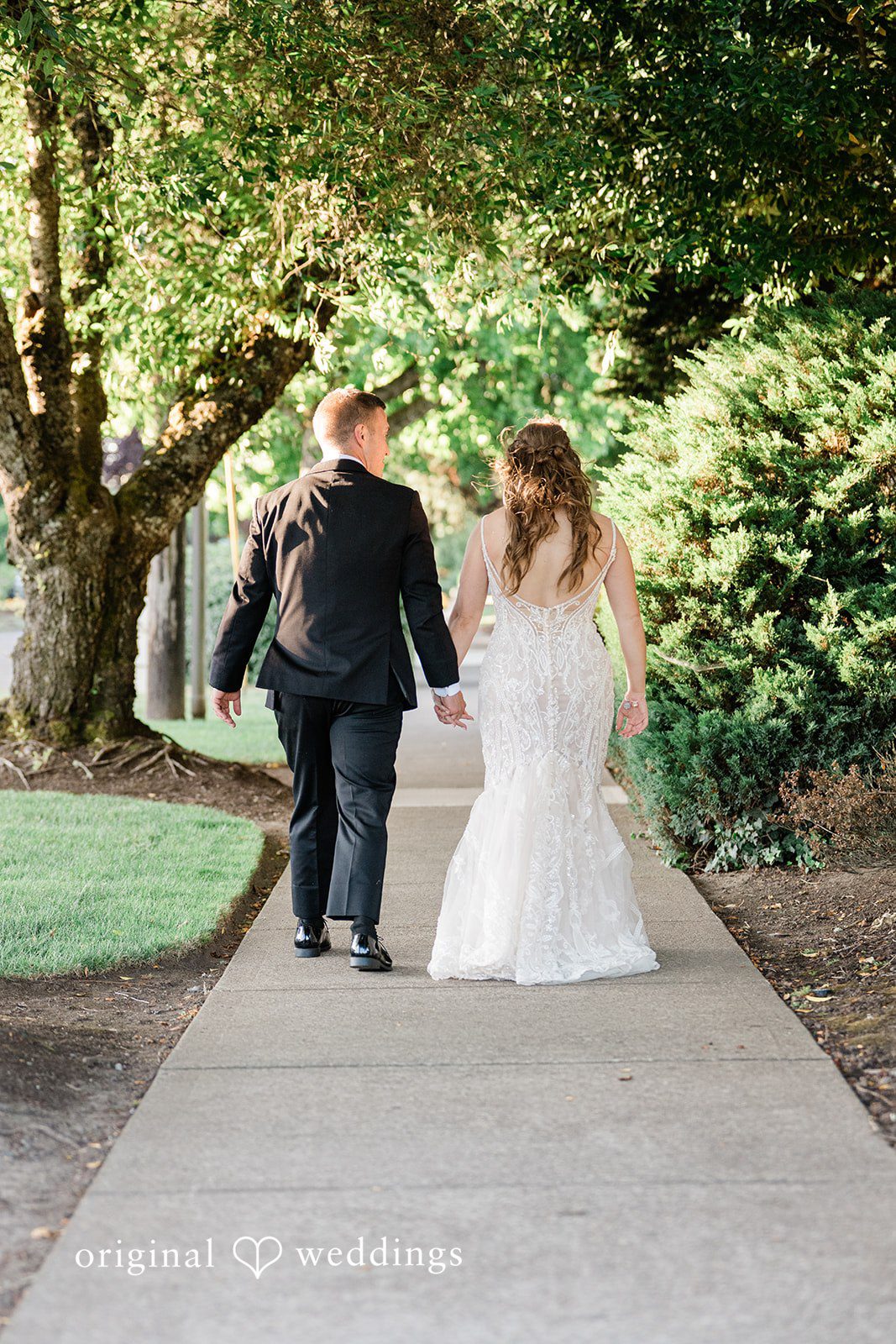 A bride and groom walking hand-in-hand down a concrete path surrounded by trees.