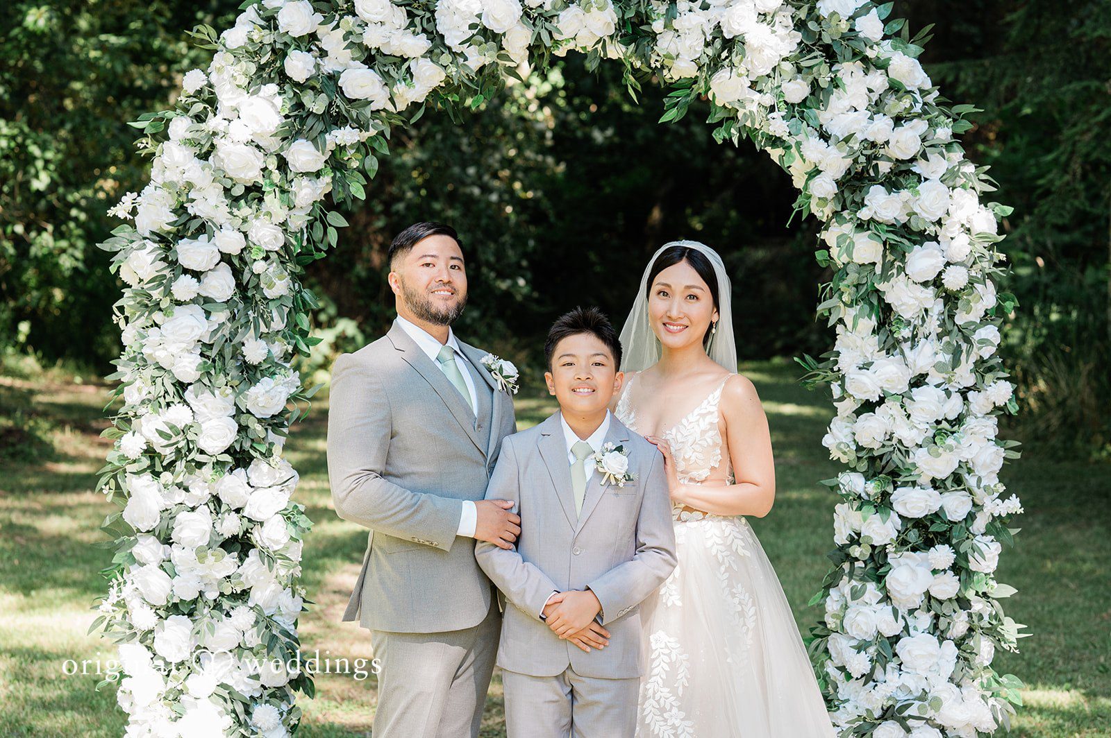 Harley + Mei A bride and groom posing with a child under a large archway of white flowers.
