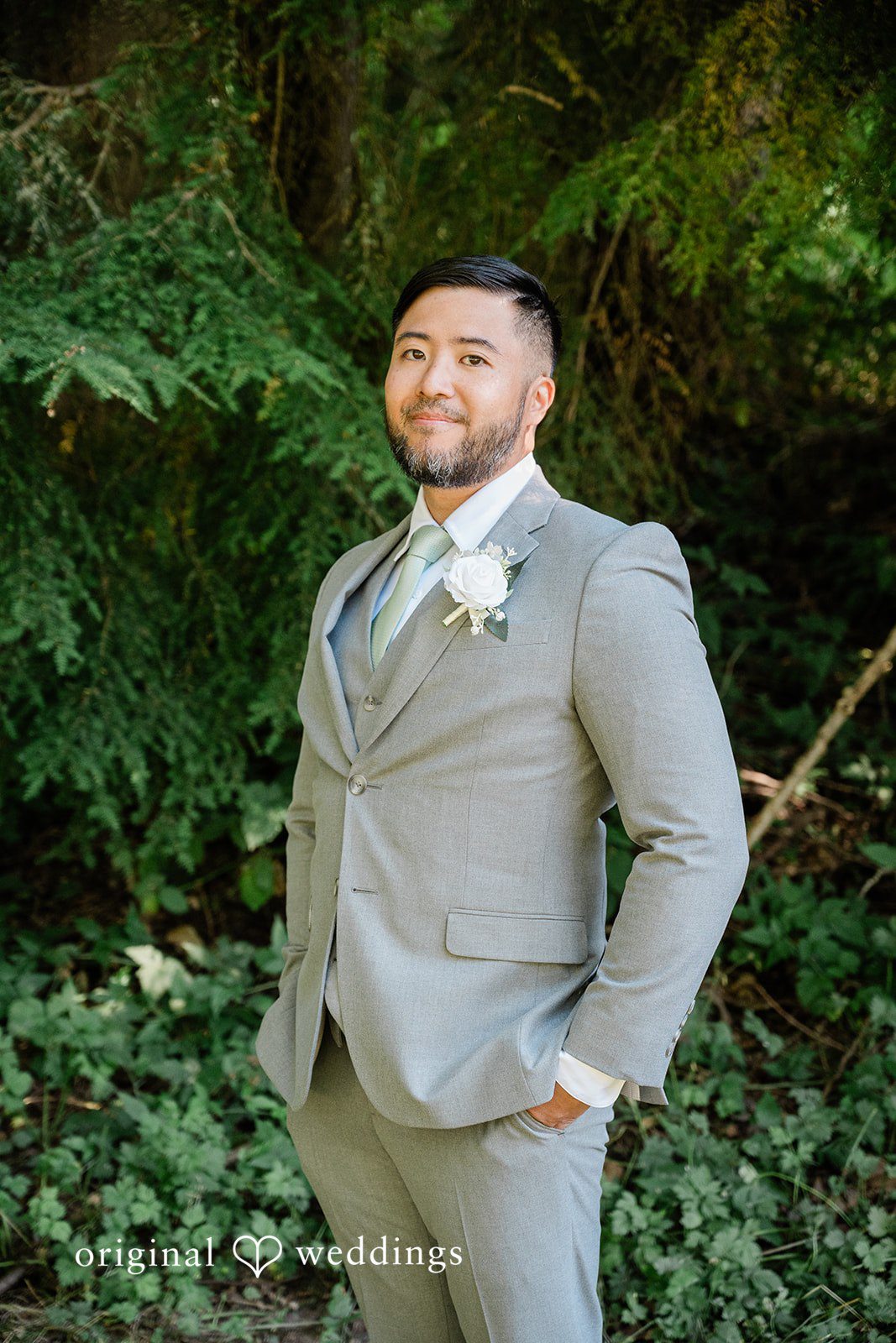 Harley + Mei A groom wearing a light grey suit smiling while standing in a wooded area.