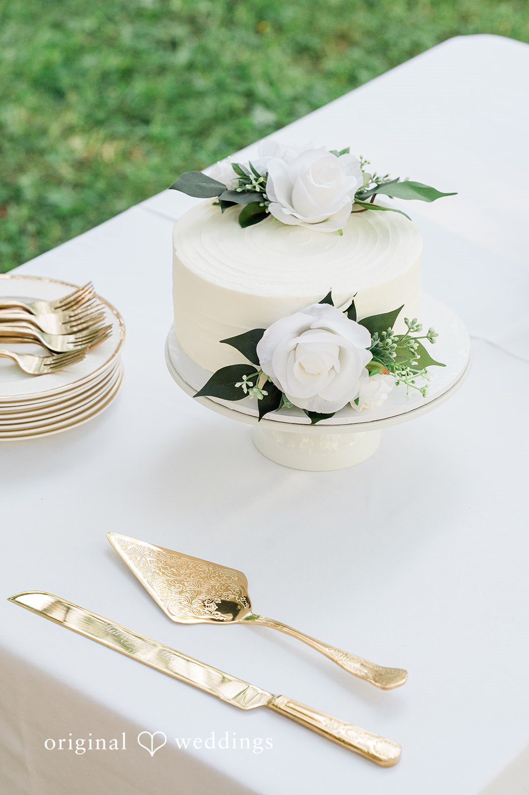 Harley + Mei A white wedding cake decorated with roses next to golden serving utensils.