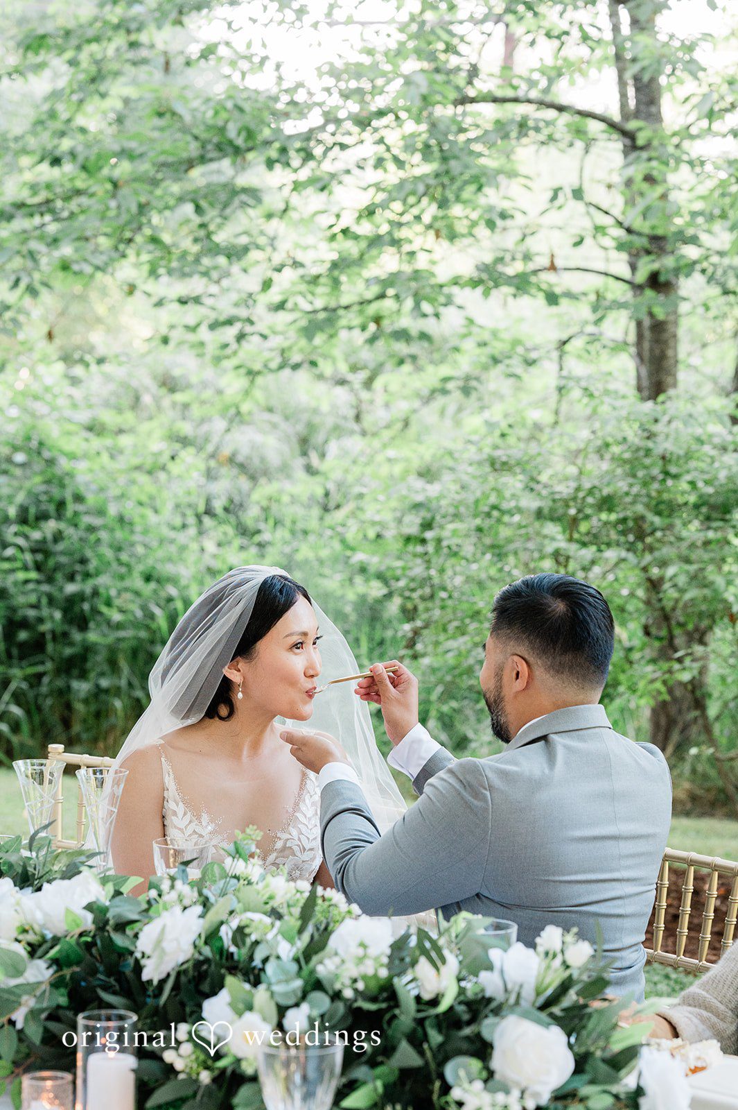 Harley + Mei A bride and groom sitting at a table looking at each other affectionately.