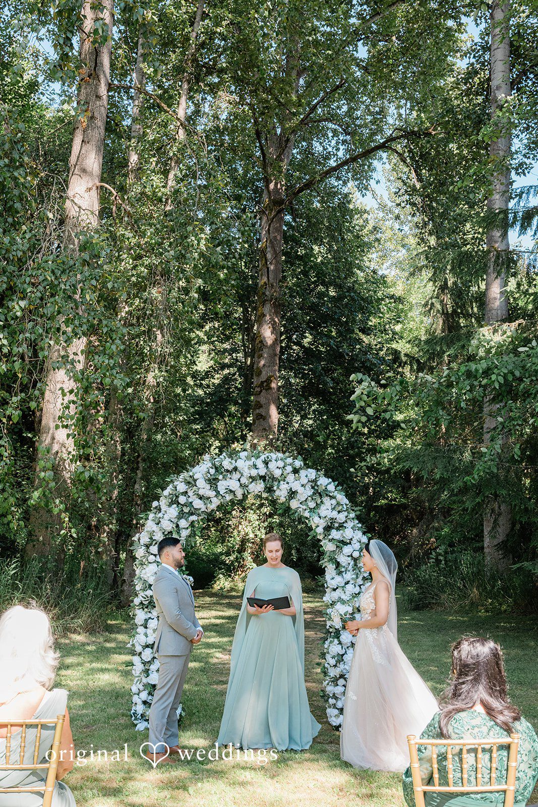 Harley + Mei A distant wedding ceremony taking place under a large floral arch in the woods.