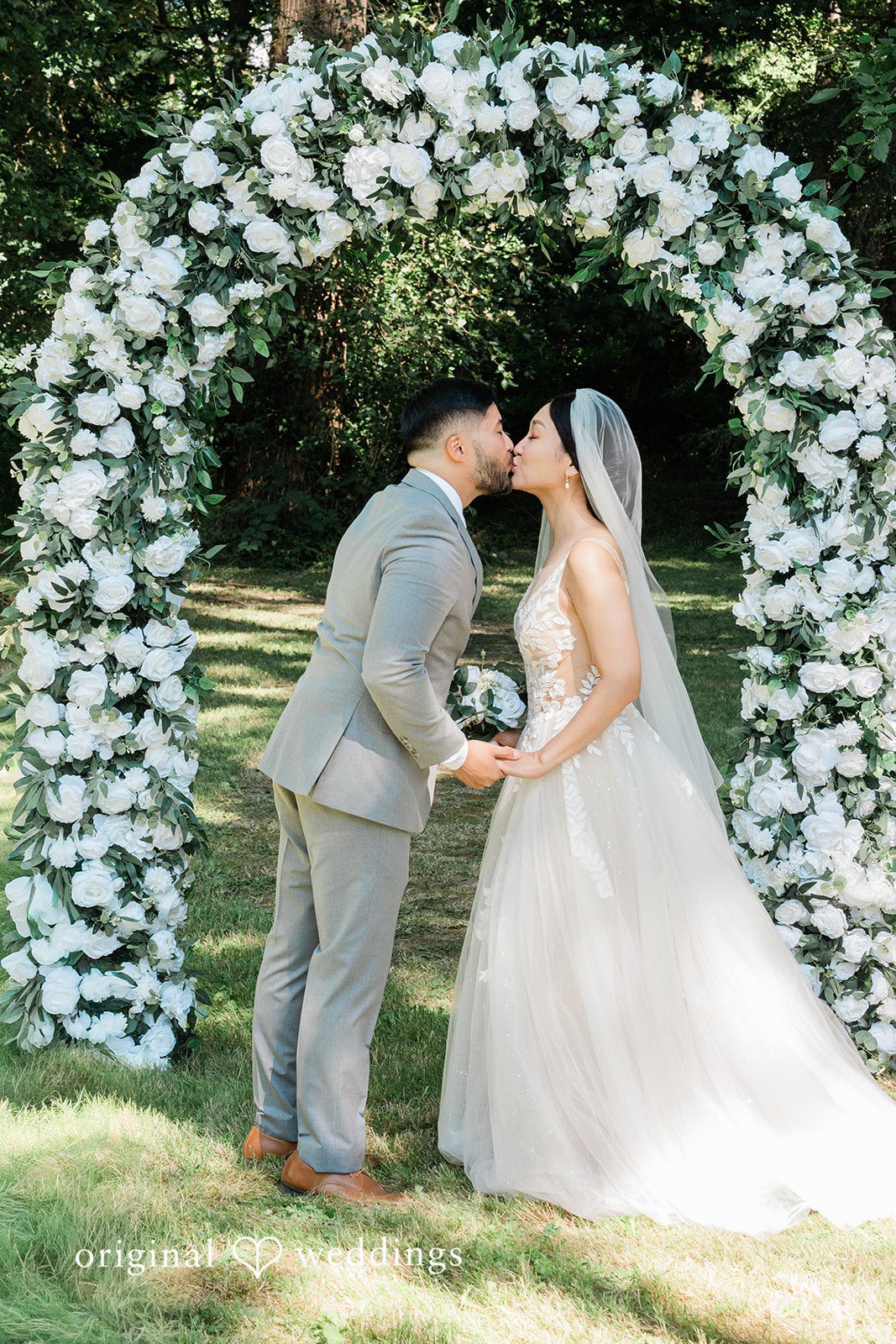 Harley + Mei A bride and groom sharing a kiss beneath a white floral archway.