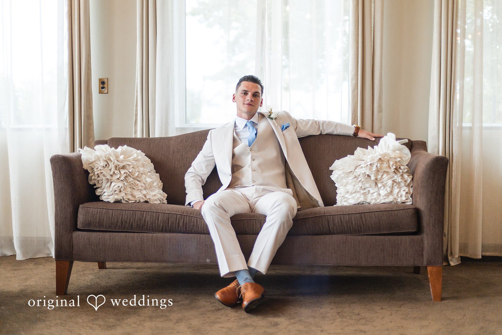 Jaclyn + Mason Groom in a light suit sitting on a tufted sofa.
