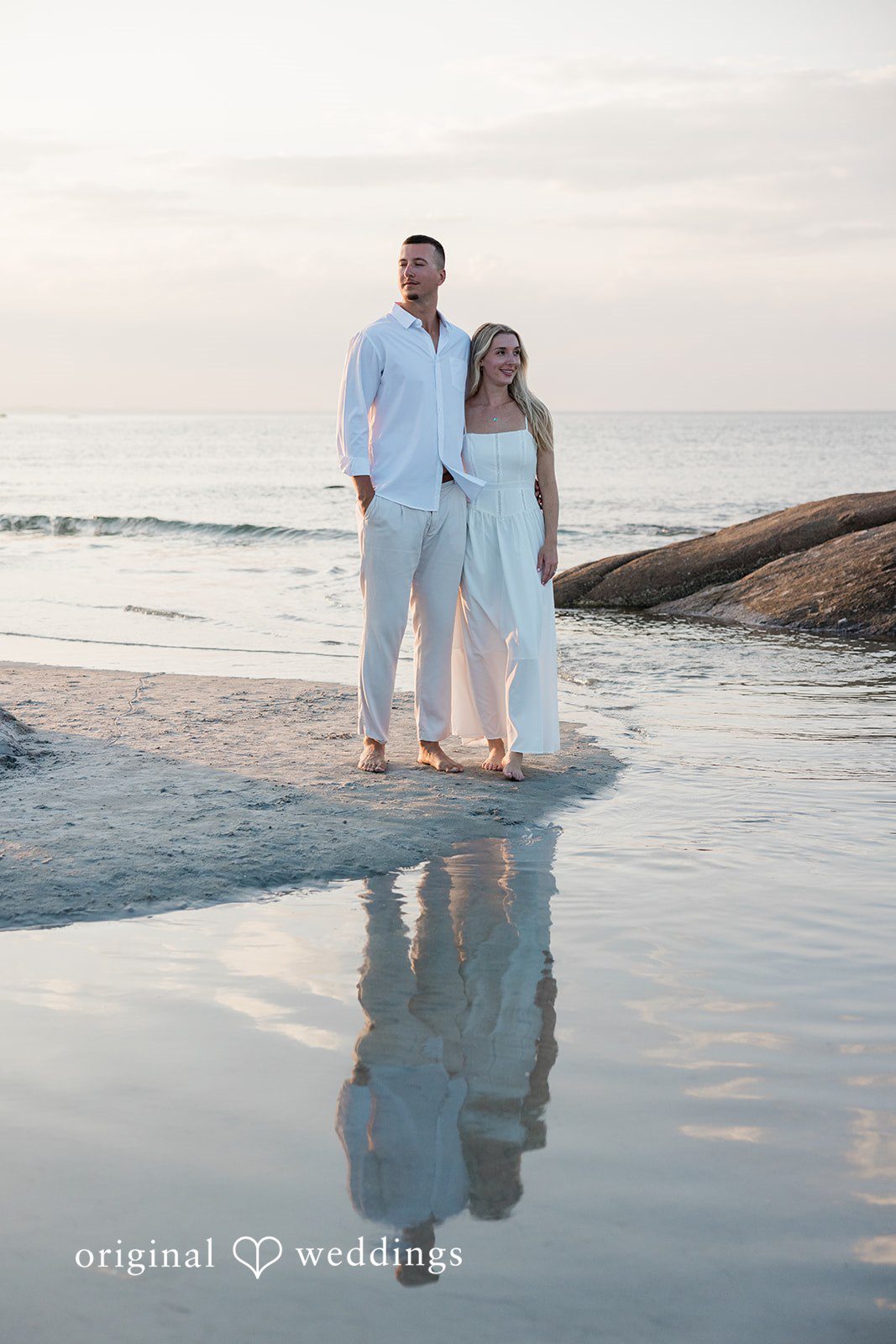 A couple standing at the edge of the water with their reflection visible on the wet sand.
