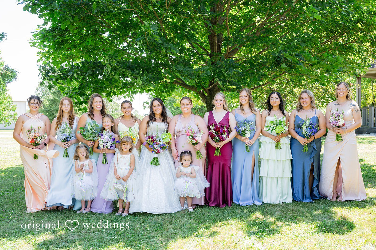 Melayna + Harley A bridal party in mismatched dresses poses together under trees.