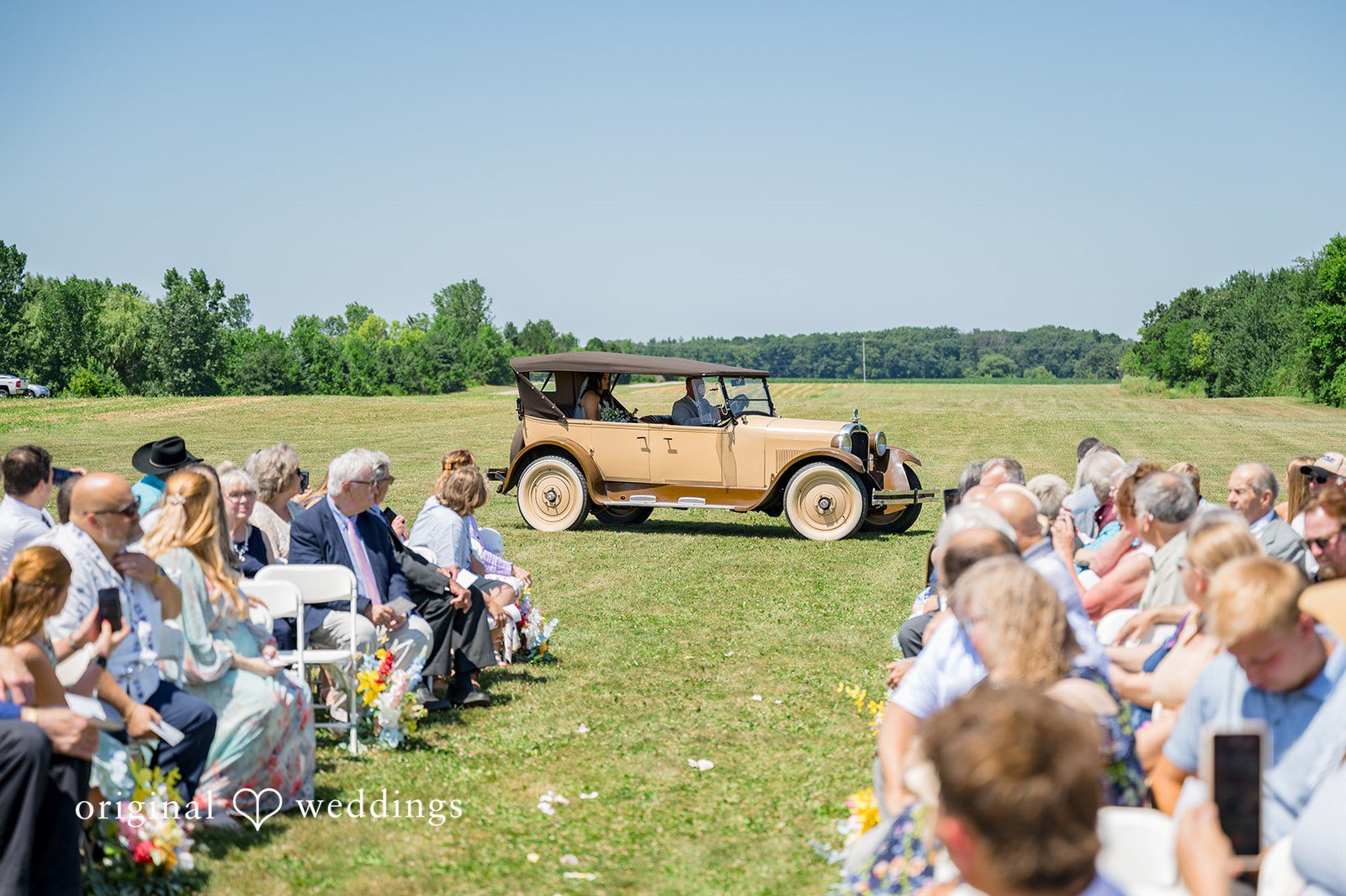 Melayna + Harley A vintage car is parked near an open-air wedding ceremony setup.