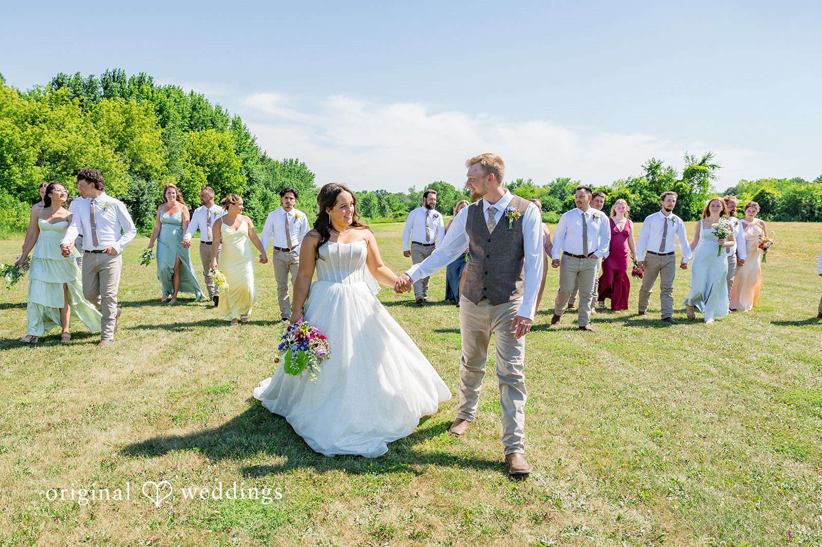 Melayna + Harley A bride and groom walk hand-in-hand through a grassy field.