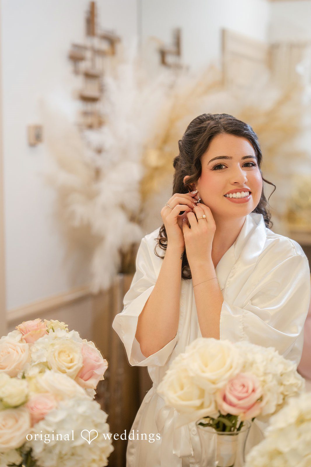 Emily + Angelo Bride in a white robe smiling while adjusting her earring before a floral backdrop.
