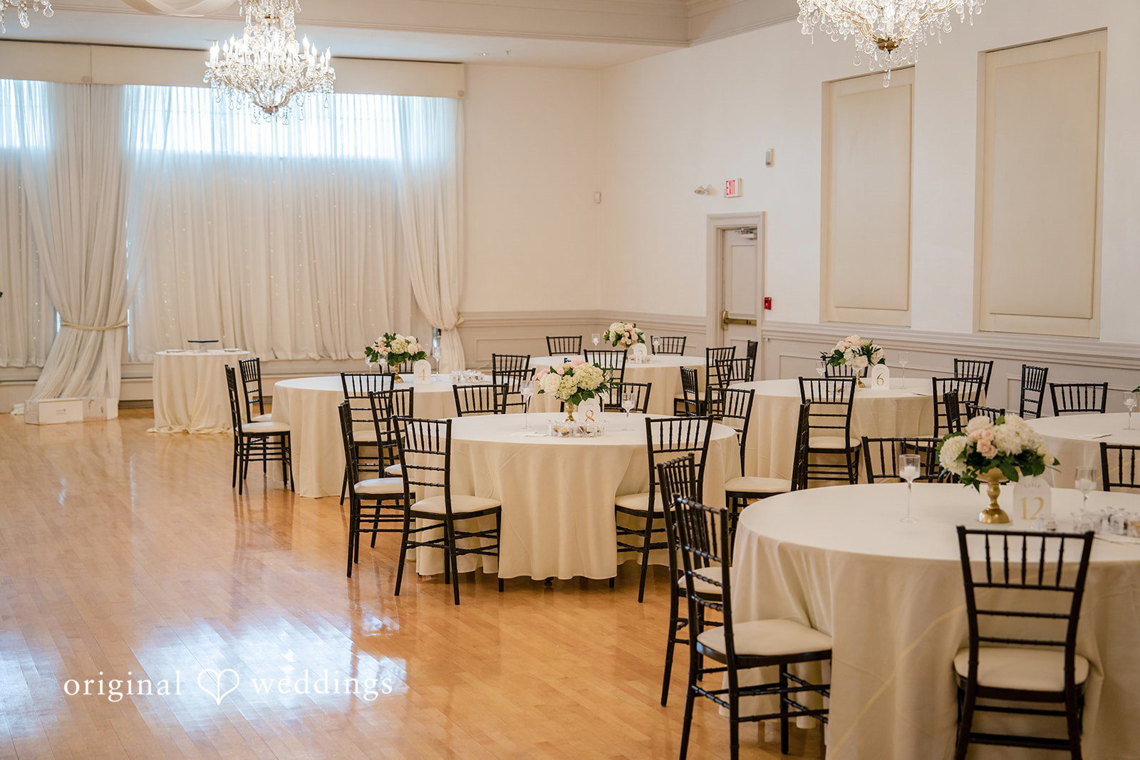 Emily + Angelo Empty reception hall arranged with round tables, white tablecloths, and dark chairs.