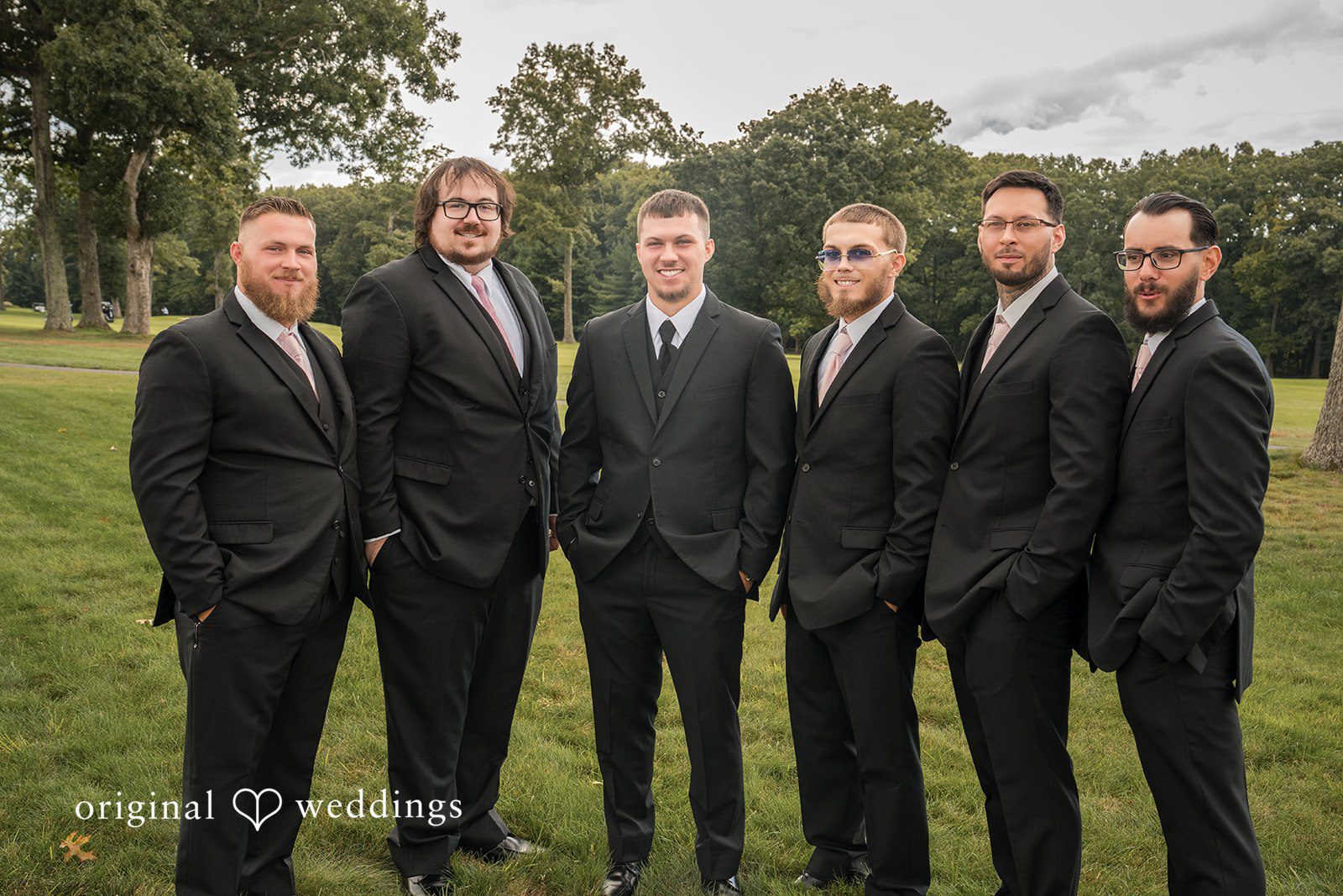 Emily + Angelo Groomsmen dressed in identical dark suits posing together in front of greenery.