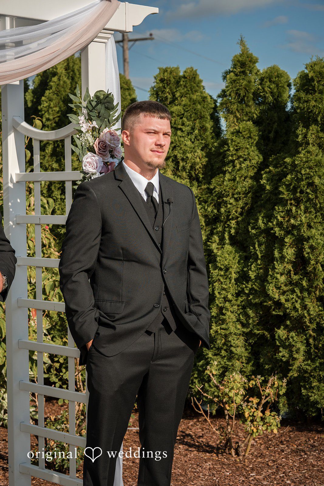 Emily + Angelo Groom in a black suit standing alone against a backdrop of green hedges.