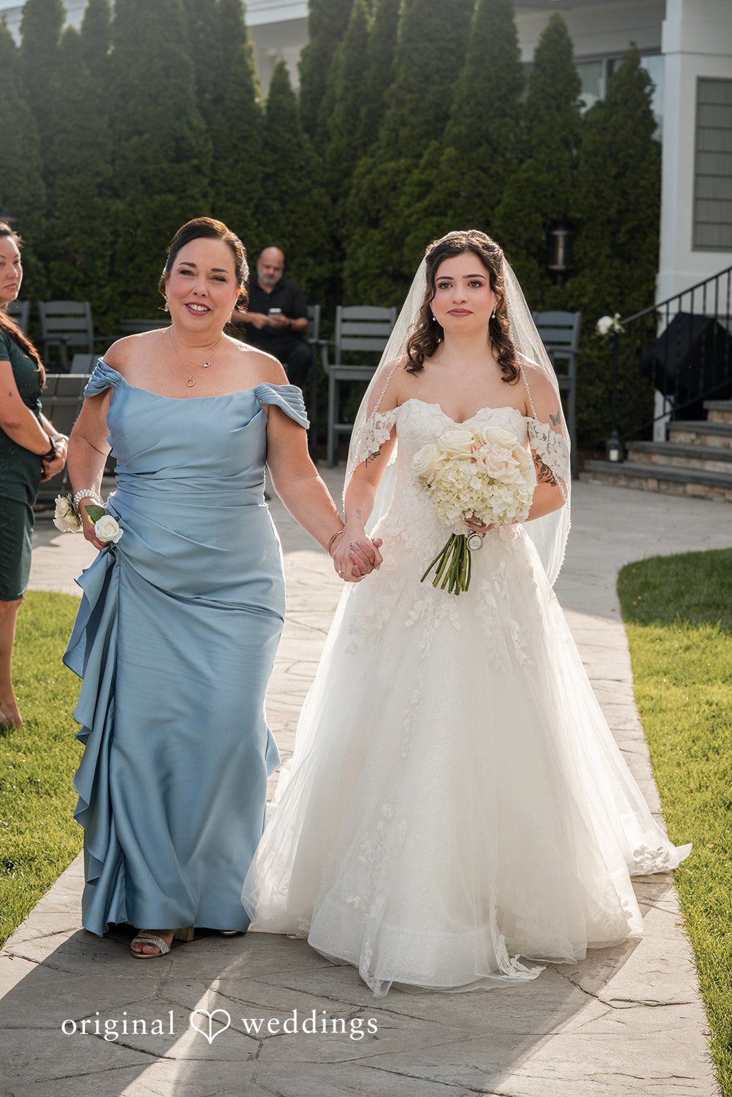 Emily + Angelo Bride and women in formal gowns walking side-by-side along a stone pathway.