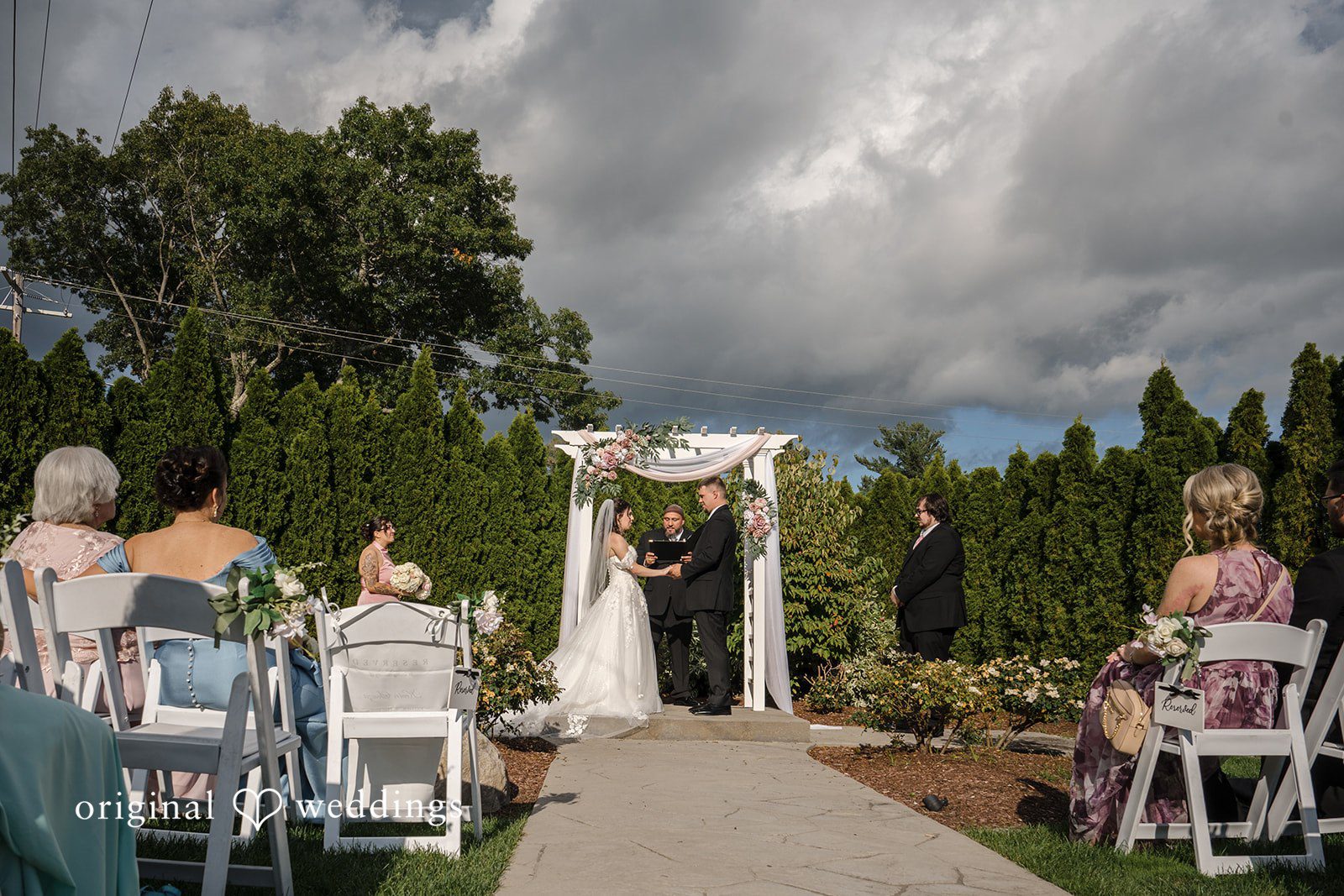 Emily + Angelo Wedding ceremony setting under a stormy gray sky with guests seated on the grass.