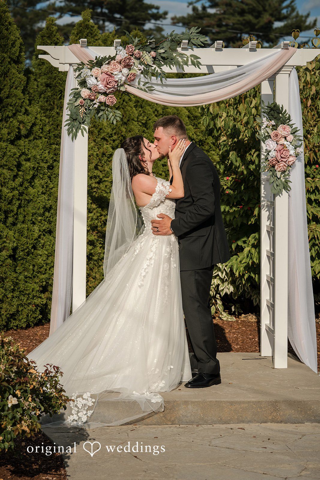 Emily + Angelo Bride and groom kissing under a white floral wedding arch during their ceremony.