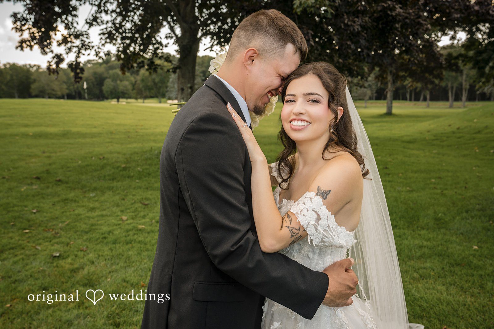 Emily + Angelo Groom embracing a smiling bride under a veil with green trees behind them.