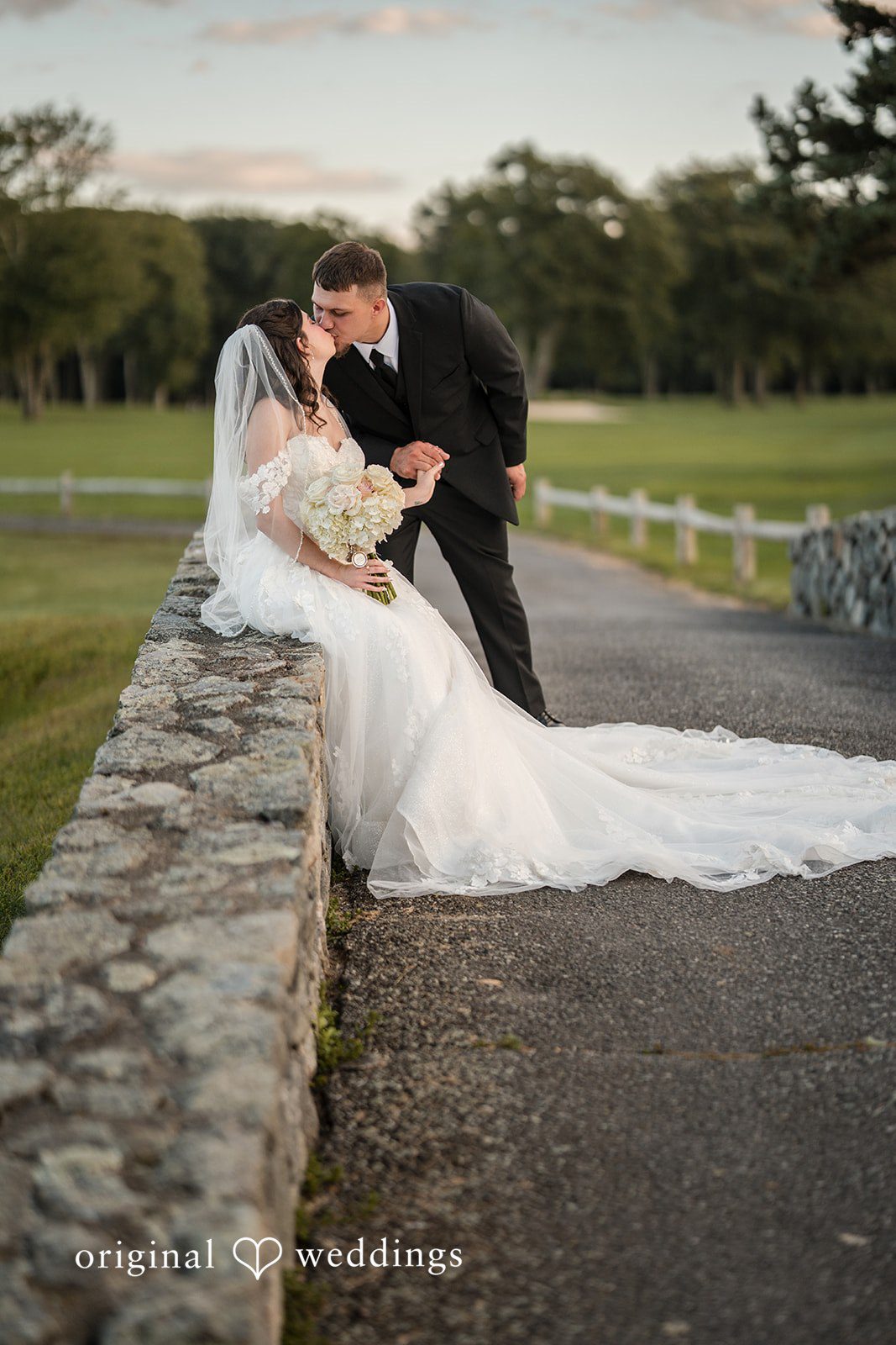 Emily + Angelo Bride and groom sitting together on a stone wall in a grassy field.