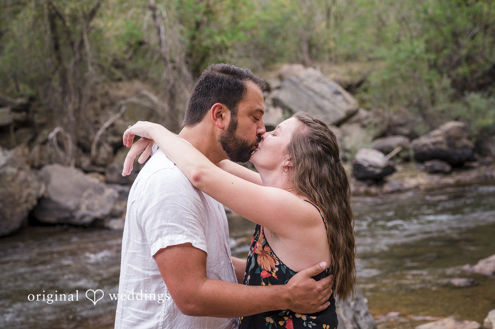 Theodore + Kathryn A man in a white shirt wrapping his arms around a woman in a dark dress next to a rocky river.