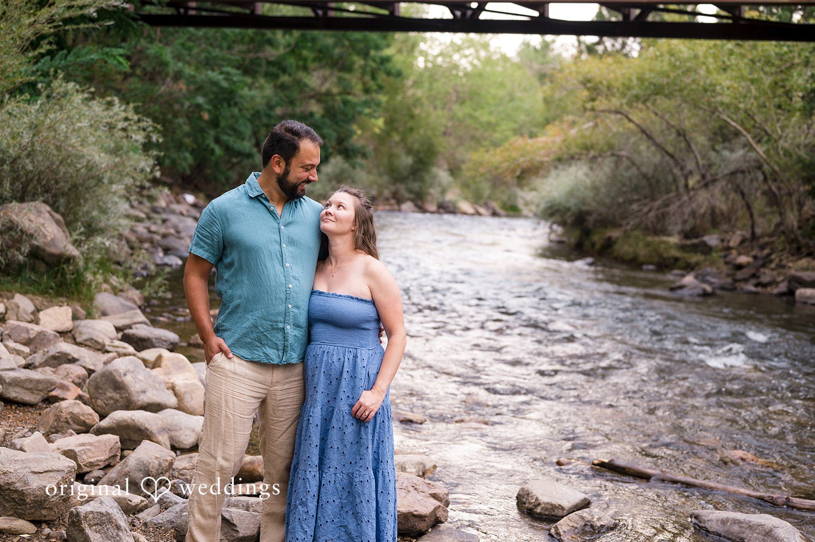 Theodore + Kathryn A man in a blue shirt and a woman in a long blue dress walking along a shallow, rocky stream.