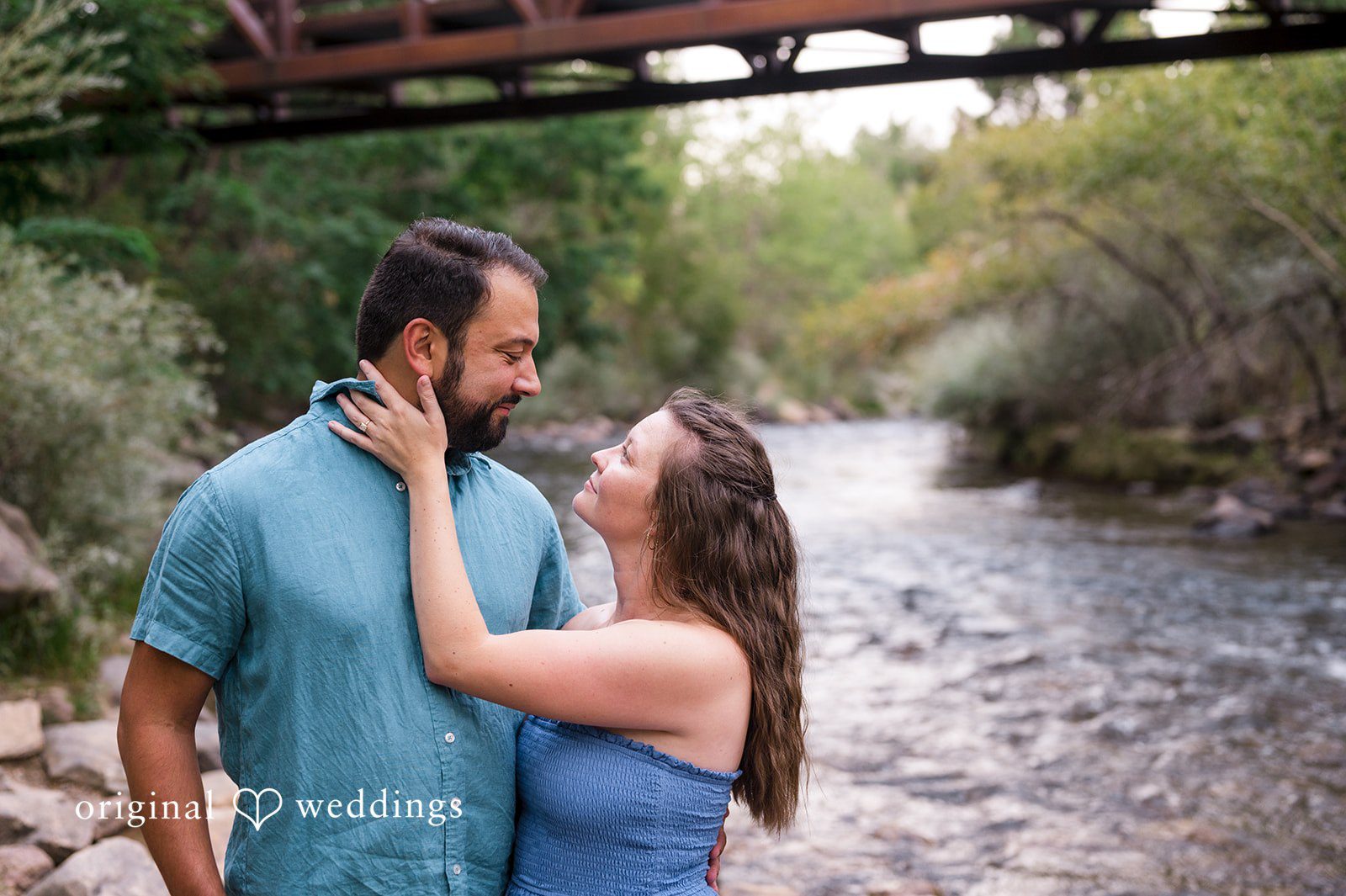 Theodore + Kathryn A man in a blue shirt and a woman in a grey dress embracing underneath a bridge structure.