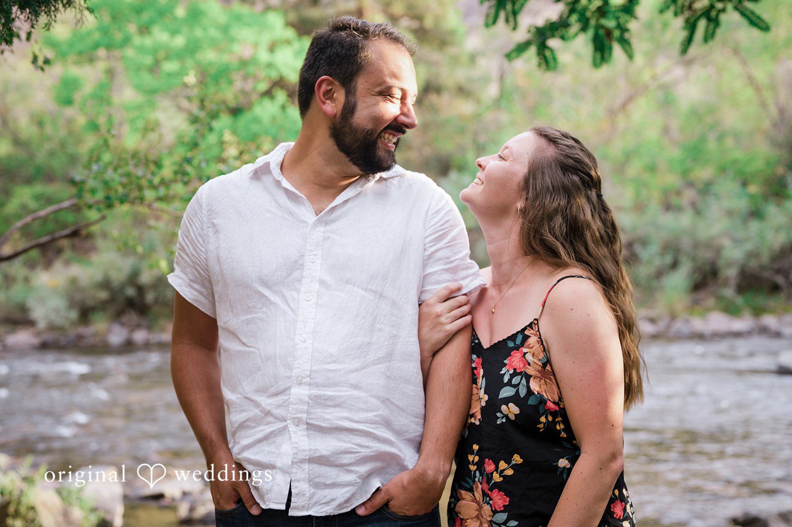 Theodore + Kathryn A man in a white shirt and a woman in a patterned dress looking lovingly at each other in a wooded area.