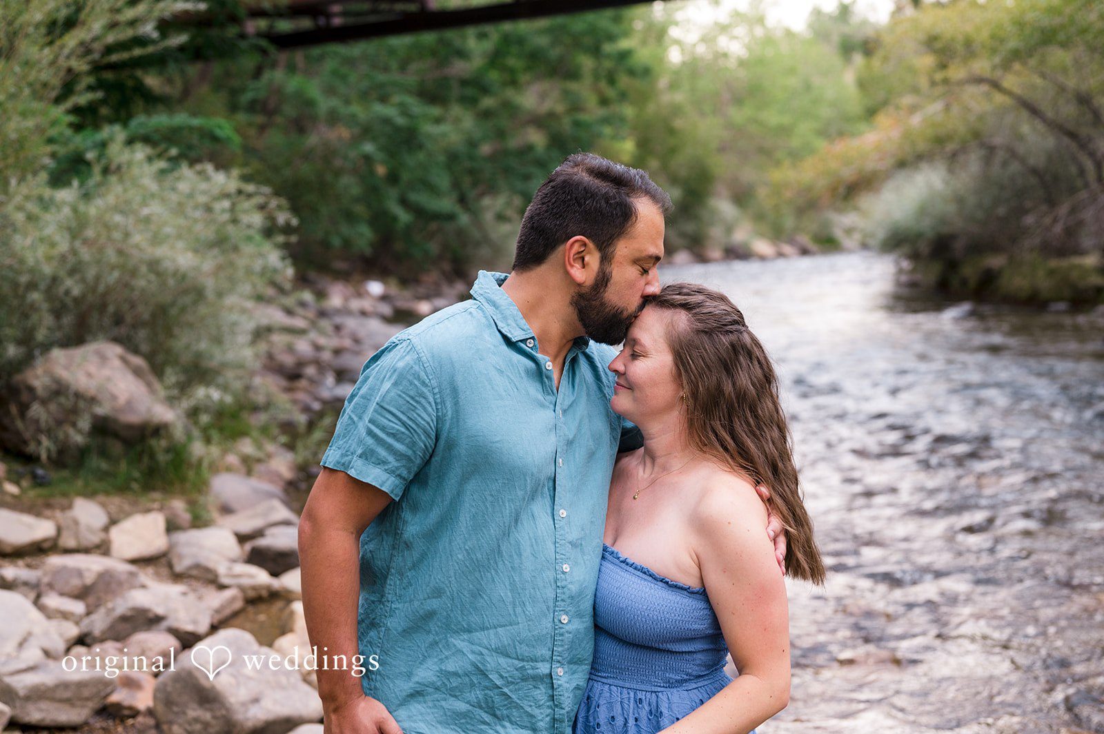 Theodore + Kathryn A man in a blue button-down shirt kissing the forehead of a woman in a grey dress by a stream.