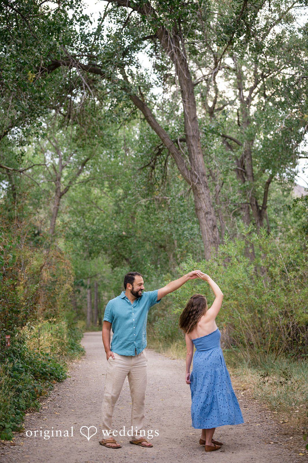 Theodore + Kathryn A man in a tan shirt and a woman in a blue dress dancing joyfully in a grassy opening surrounded by trees.