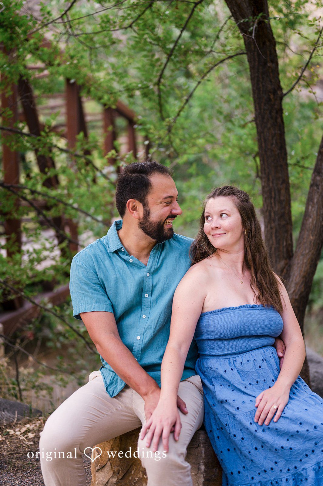 Theodore + Kathryn A man in a blue shirt and a woman in a strapless blue dress sitting together on the ground in a forest.
