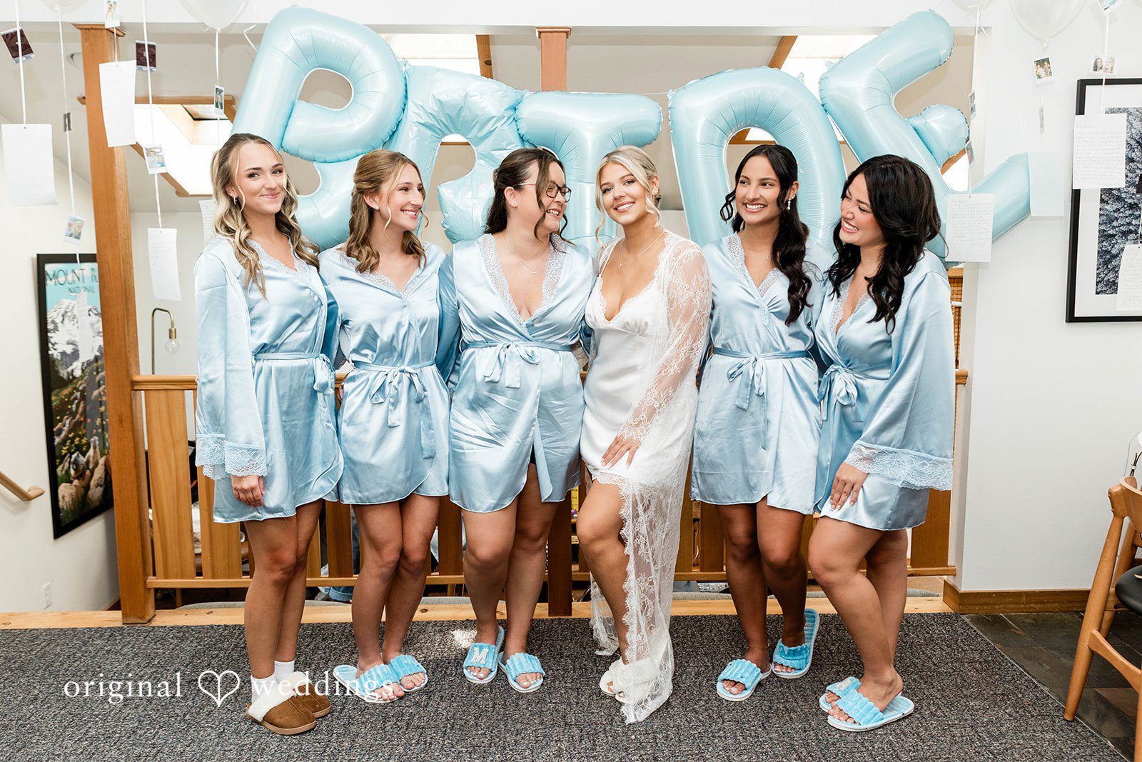 Bethany + Ryan A group of women in matching light-blue satin robes pose together on a bed.