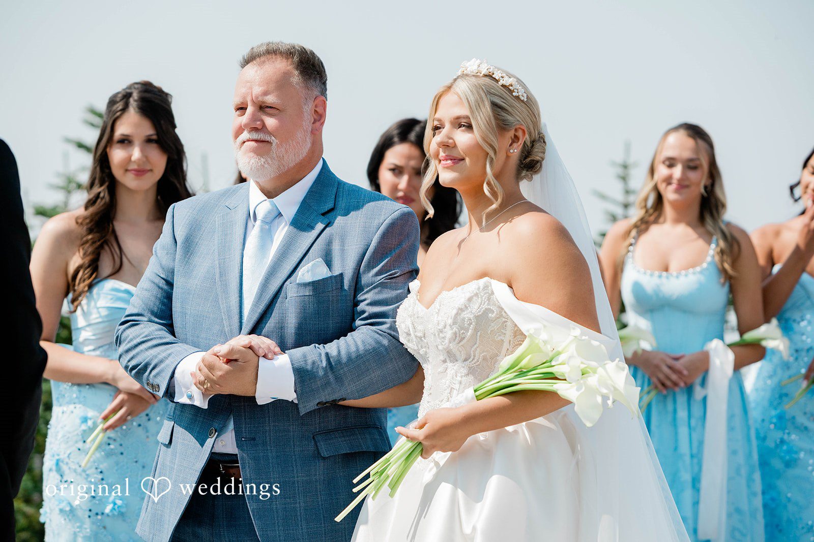 Bethany + Ryan A bride in a white strapless gown holds a bouquet of white calla lilies while walking arm-in-arm with a man in a blue suit.