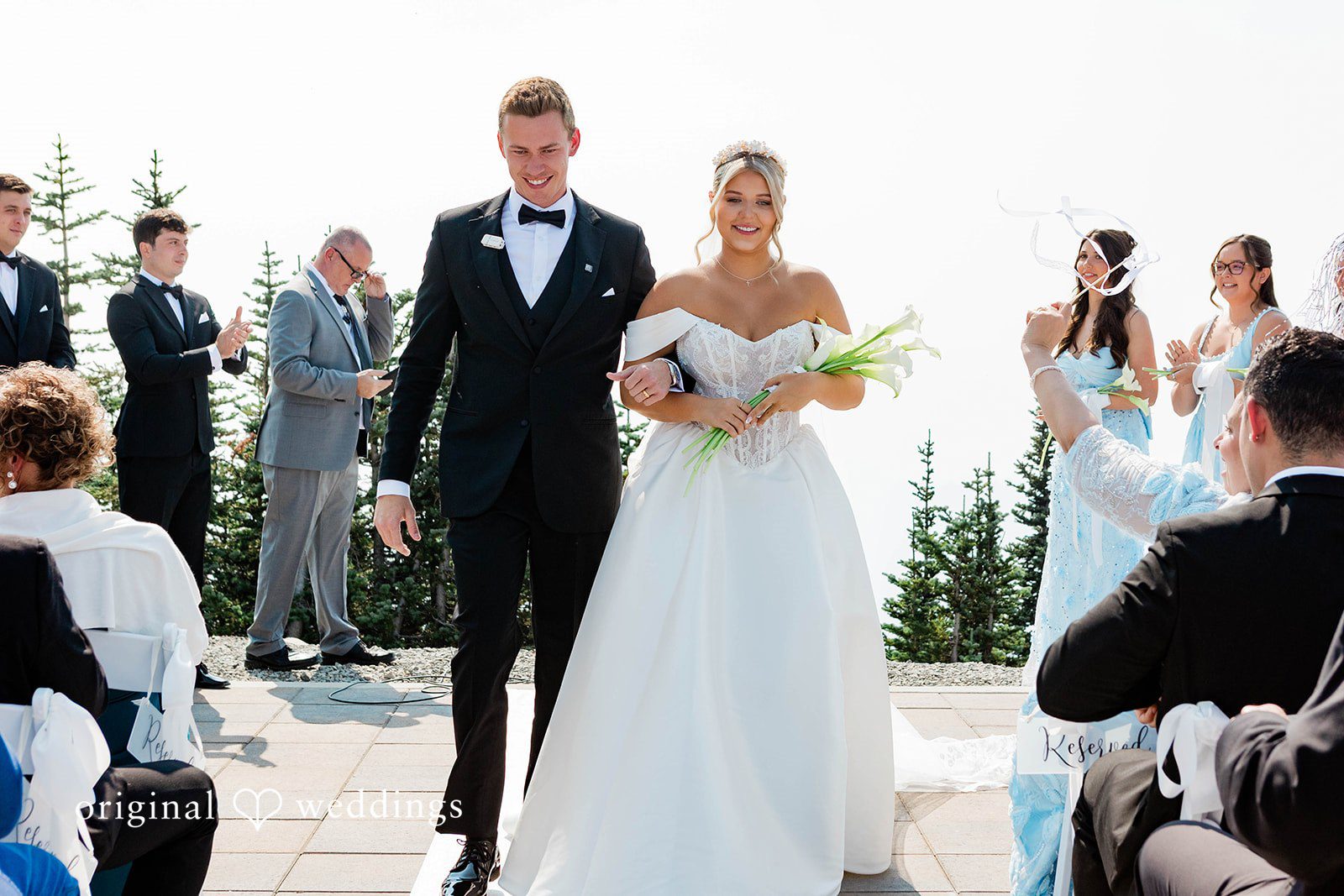 Bethany + Ryan A bride and groom walk hand-in-hand down the aisle while guests applaud.